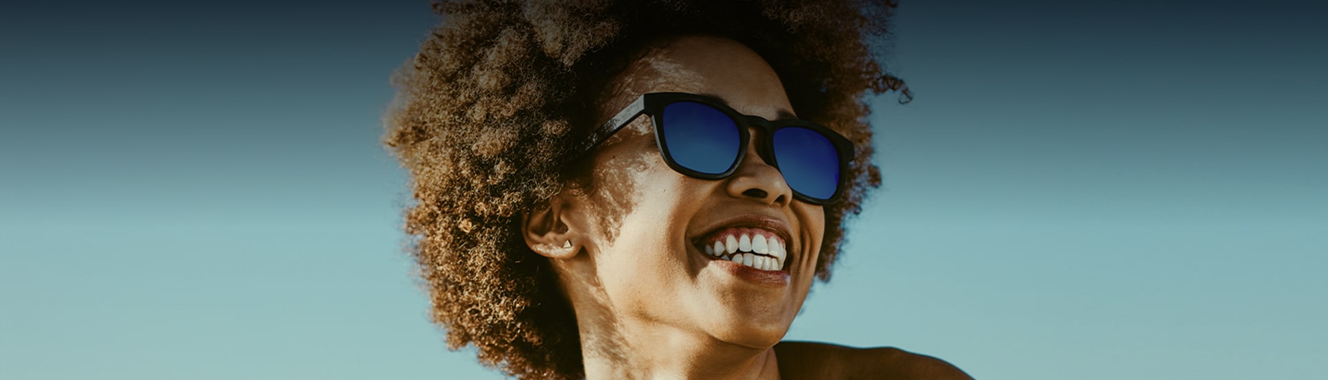 woman with curly hair smiling and wearing sunglasses against a blue sky